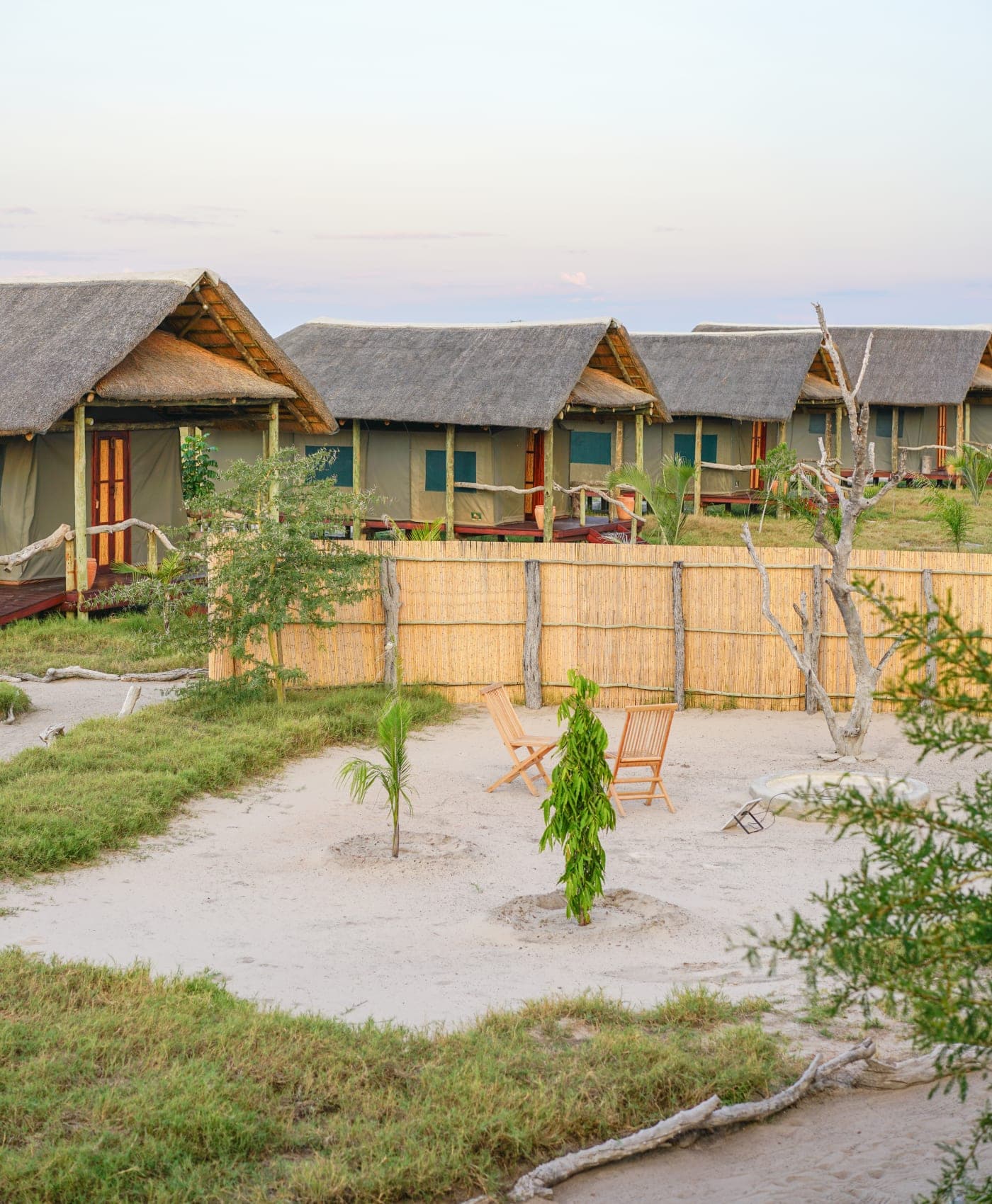 Une rangée de chambres sous tente et toit de chaume au crépuscule, chacune avec véranda et chaises longues sur le sable