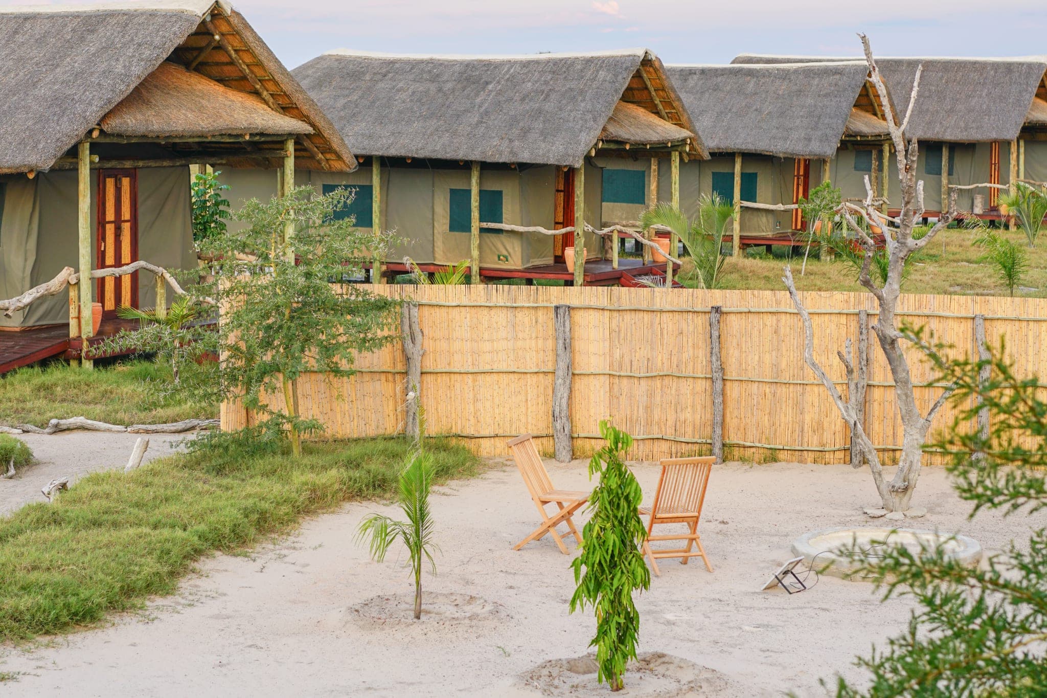 Une rangée de chambres sous tente et toit de chaume au crépuscule, chacune avec véranda et chaises longues sur le sable